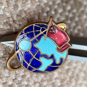 Può includere: Uno spillo in smalto blu e oro con un design a globo e una bandiera rossa con una falce e un martello. Il globo è circondato da un anello dorato.