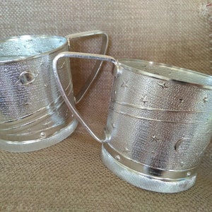 Puede incluir: Dos tazas de metal plateado con asas. Las tazas tienen una superficie texturizada y están decoradas con un patrón de estrellas y una luna creciente.