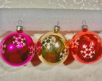 May include: Three vintage glass Christmas ornaments with snowflake designs. The ornaments are pink, gold, and red.