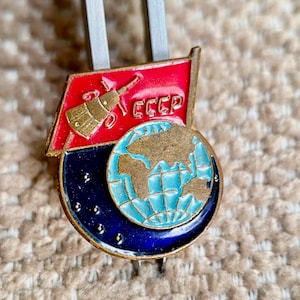May include: A vintage enamel pin featuring a red flag with a rocket and the text "CCCP" above a blue globe with a gold outline. The globe is surrounded by a ring of gold stars.