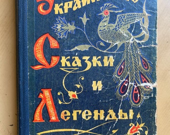 Puede incluir: Un libro azul con el título "Українські казки і легенди" en letras doradas. La portada presenta un pavo real estilizado con acentos dorados y rojos.