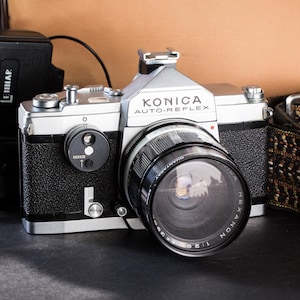 May include: A vintage Konica Auto-Reflex camera with a black and silver body, a large lens, and a flash unit. A yellow film canister with a green lid and a brown leather bag are also present. The camera is a classic photography item.