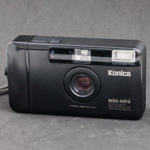 May include: A black Konica Big Mini point-and-shoot camera featuring a 35mm F3.5 lens, auto-variable flash, auto-close up, red-eye reduction, and auto-focus capabilities. The front of the camera displays the Konica brand name and model text.