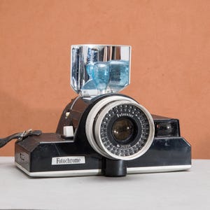 May include: A black Fotochrome camera with a silver and black lens ring featuring the text Fotocolor and No. 202239. The camera includes a top-mounted chrome flash reflector with a blue bulb and a black carrying strap attached to the side.