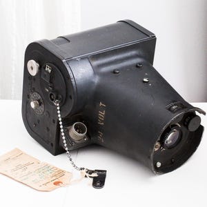 May include: Black vintage camera with a lens and various dials. The camera has the text "24 VOLT" printed on the side. A tag is attached to the camera with a chain.
