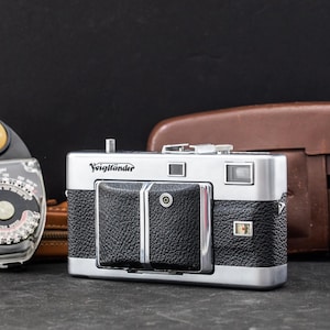 May include: A silver and black Voigtländer camera with a textured grip, a central lens cover, and a viewfinder window. Beside it sits a black light meter with a yellow dome and a brown leather camera case positioned in the background.