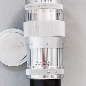 Puede incluir: Un objetivo de c&aacute;mara Canon plateado con base negra y agarre blanco acanalado. El objetivo tiene marcas de medici&oacute;n y una tapa con el logotipo de Canon. Dise&ntilde;ado para fotograf&iacute;a.