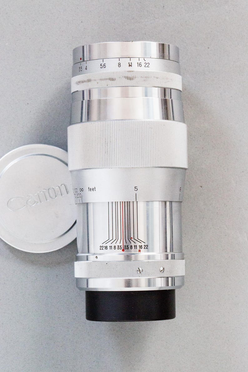 Puede incluir: Un objetivo de c&aacute;mara Canon plateado con base negra y agarre blanco acanalado. El objetivo tiene marcas de medici&oacute;n y una tapa con el logotipo de Canon. Dise&ntilde;ado para fotograf&iacute;a.