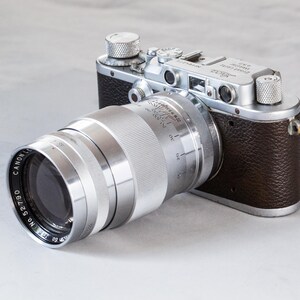 Puede incluir: Una c&aacute;mara vintage con una lente plateada y un cuerpo de cuero marr&oacute;n. La lente es grande y cil&iacute;ndrica, con el texto "CANON LENS" visible. El cuerpo de la c&aacute;mara tiene diales plateados.
