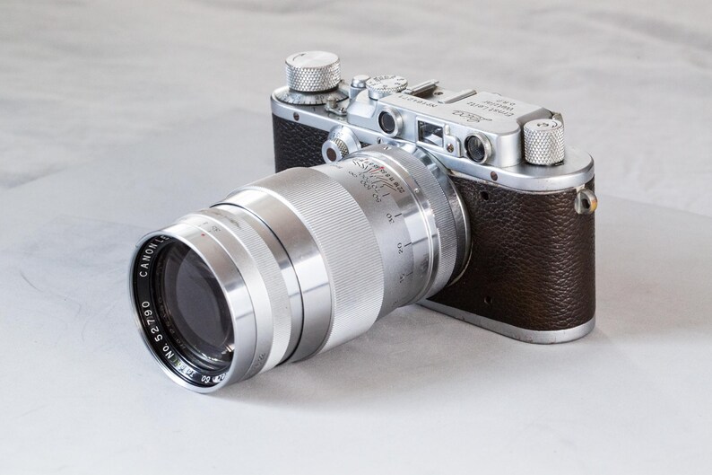 Puede incluir: Una c&aacute;mara vintage con una lente plateada y un cuerpo de cuero marr&oacute;n. La lente es grande y cil&iacute;ndrica, con el texto "CANON LENS" visible. El cuerpo de la c&aacute;mara tiene diales plateados.