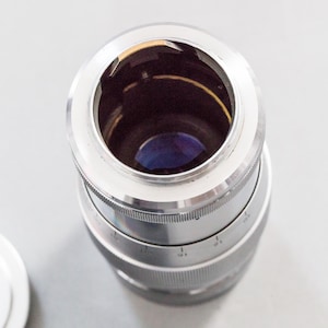 Puede incluir: Un objetivo de c&aacute;mara plateado con interior negro y lente azul. El objetivo tiene un anillo de enfoque estriado y un borde blanco. El objetivo es cil&iacute;ndrico.