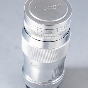 Puede incluir: Un objetivo de c&aacute;mara Canon plateado con el logotipo de Canon en la tapa del objetivo. El objetivo tiene marcas de medici&oacute;n y un anillo de enfoque. El objetivo es cil&iacute;ndrico y tiene una base negra.