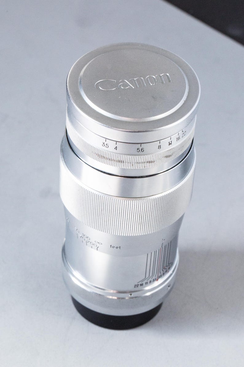 Puede incluir: Un objetivo de c&aacute;mara Canon plateado con el logotipo de Canon en la tapa del objetivo. El objetivo tiene marcas de medici&oacute;n y un anillo de enfoque. El objetivo es cil&iacute;ndrico y tiene una base negra.