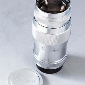 Puede incluir: Un objetivo Canon 135mm f/3.5 plateado con una tapa de objetivo a juego. El objetivo tiene un cuerpo acanalado y detalles en negro. La tapa del objetivo es redonda con el logotipo de Canon.
