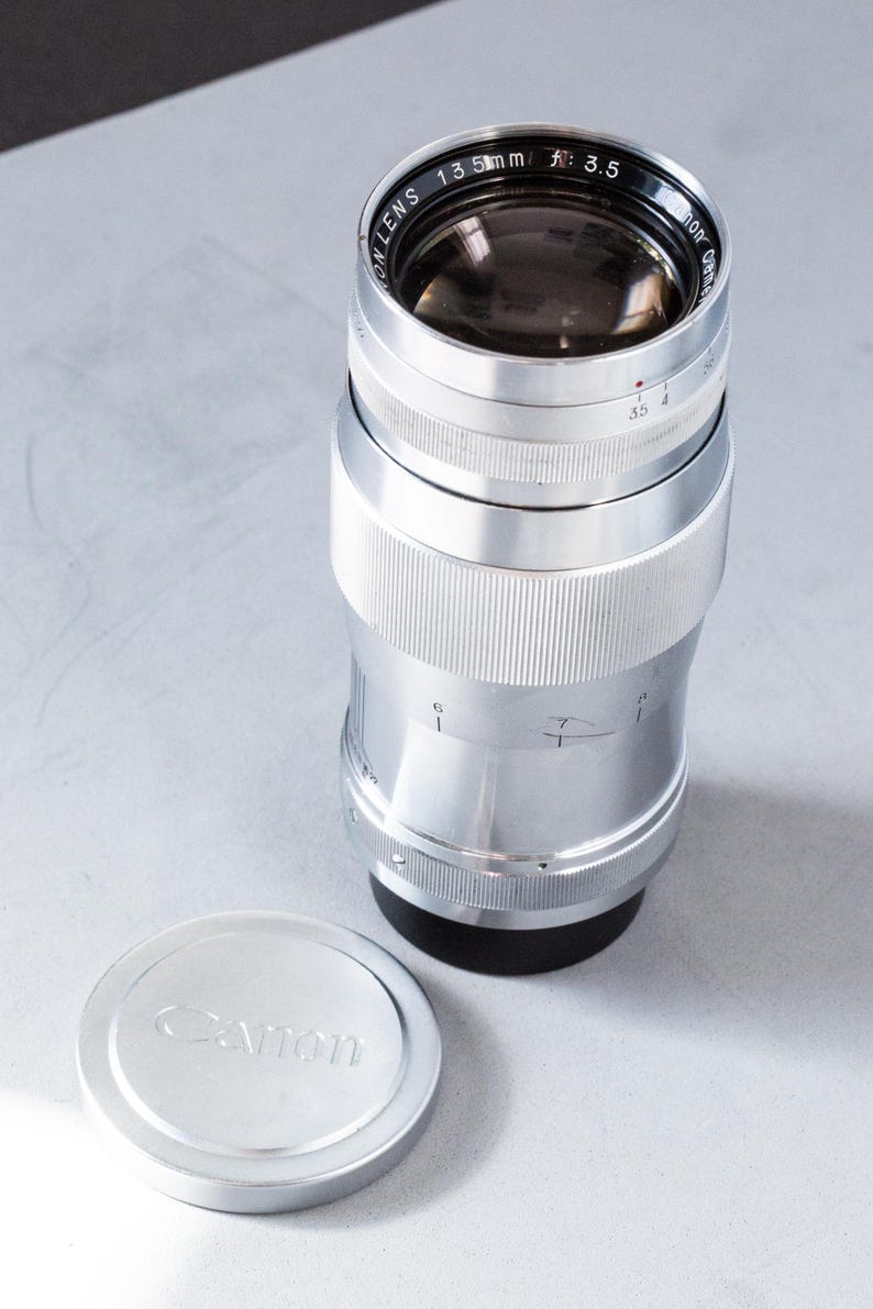 Puede incluir: Un objetivo Canon 135mm f/3.5 plateado con una tapa de objetivo a juego. El objetivo tiene un cuerpo acanalado y detalles en negro. La tapa del objetivo es redonda con el logotipo de Canon.