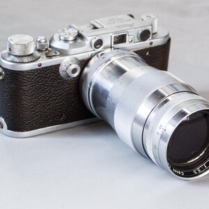 Puede incluir: Una c&aacute;mara plateada y negra vintage con una lente Canon. La lente est&aacute; marcada como "Canon Lens 135mm f/3.5". El cuerpo de la c&aacute;mara es negro con detalles plateados. La c&aacute;mara est&aacute; sobre una superficie gris claro.