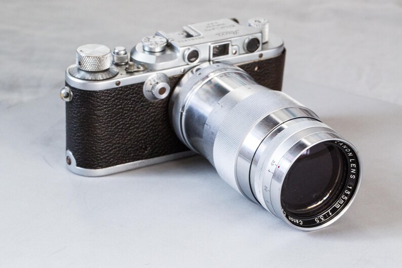 Puede incluir: Una c&aacute;mara plateada y negra vintage con una lente Canon. La lente est&aacute; marcada como "Canon Lens 135mm f/3.5". El cuerpo de la c&aacute;mara es negro con detalles plateados. La c&aacute;mara est&aacute; sobre una superficie gris claro.