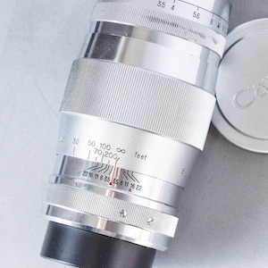 Puede incluir: Lente de c&aacute;mara plateada con detalles en negro y una tapa de lente. La lente tiene marcas de medici&oacute;n en pies y una gama de diafragmas. La lente est&aacute; dise&ntilde;ada para una distancia focal de 70-200 mm.