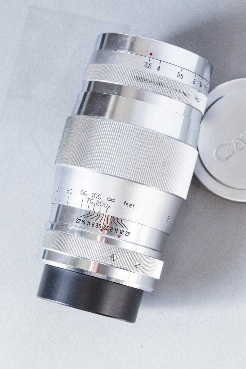 Puede incluir: Lente de c&aacute;mara plateada con detalles en negro y una tapa de lente. La lente tiene marcas de medici&oacute;n en pies y una gama de diafragmas. La lente est&aacute; dise&ntilde;ada para una distancia focal de 70-200 mm.
