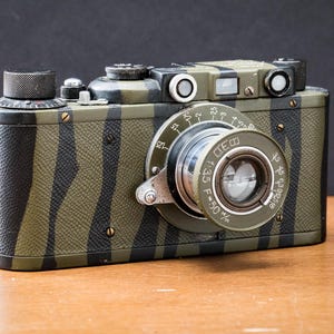May include: Vintage camera with a camouflage pattern of black and olive green stripes. The lens is silver and has a circular dial with numbers. The camera is on a wooden surface with a dark background.