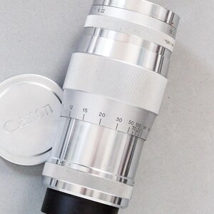 Puede incluir: Un objetivo de c&aacute;mara Canon plateado con una base negra y una tapa de objetivo a juego. El objetivo tiene marcas de medici&oacute;n y un agarre estriado. La tapa tiene el logotipo de Canon. El objetivo est&aacute; inclinado sobre una superficie gris claro.