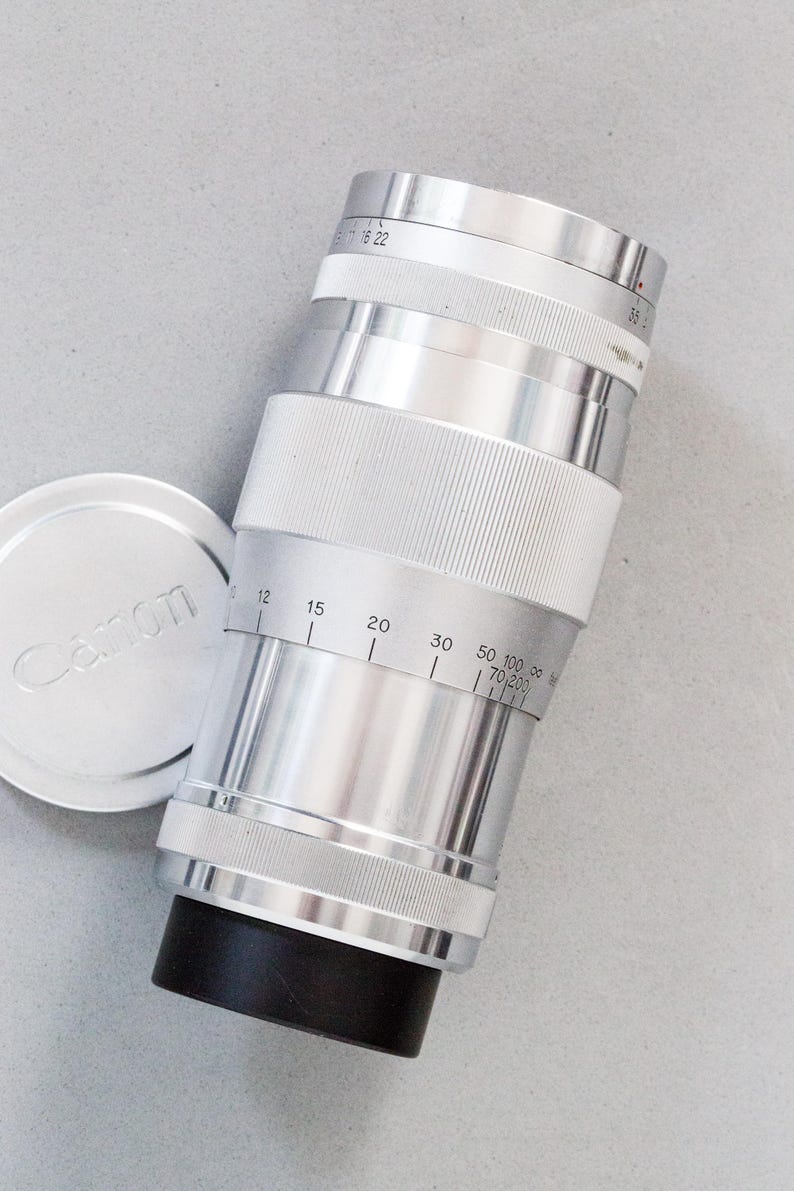 Puede incluir: Un objetivo de c&aacute;mara Canon plateado con una base negra y una tapa de objetivo a juego. El objetivo tiene marcas de medici&oacute;n y un agarre estriado. La tapa tiene el logotipo de Canon. El objetivo est&aacute; inclinado sobre una superficie gris claro.
