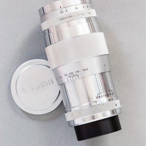 Puede incluir: Un objetivo de c&aacute;mara Canon plateado con base negra y detalles blancos. El objetivo tiene marcas de medici&oacute;n y una tapa de objetivo separada con el logotipo de Canon. El objetivo es cil&iacute;ndrico.