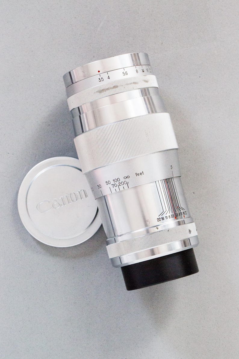 Puede incluir: Un objetivo de c&aacute;mara Canon plateado con base negra y detalles blancos. El objetivo tiene marcas de medici&oacute;n y una tapa de objetivo separada con el logotipo de Canon. El objetivo es cil&iacute;ndrico.
