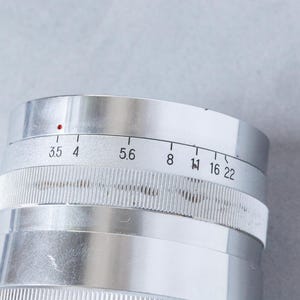 Puede incluir: Primer plano de una lente de c&aacute;mara plateada con ajustes de apertura. La lente tiene un anillo de enfoque estriado y una serie de n&uacute;meros que indican los valores de apertura, incluyendo 3.5, 4, 5.6, 8, 11, 16 y 22. Un punto rojo es visible.