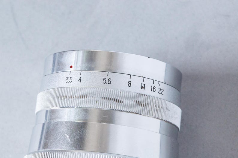 Puede incluir: Primer plano de una lente de c&aacute;mara plateada con ajustes de apertura. La lente tiene un anillo de enfoque estriado y una serie de n&uacute;meros que indican los valores de apertura, incluyendo 3.5, 4, 5.6, 8, 11, 16 y 22. Un punto rojo es visible.