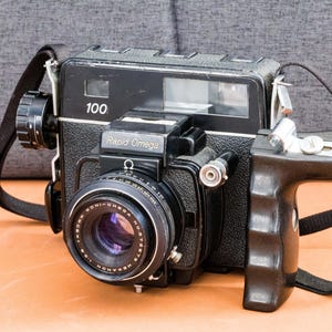 May include: Black Rapid Omega 100 medium format camera with a black strap and handle. The camera has a lens with the text "KONI-OMEGA" and "1:3.5 f=75mm". The camera has the number "100" printed on the top.