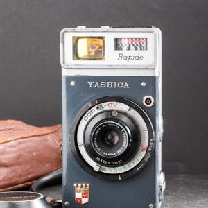 May include: A vintage Yashica Rapide camera in a gray and silver finish. The camera has a black lens with white markings and a red and white dial. The camera is accompanied by a brown leather case.