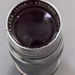 Puede incluir: Un objetivo de c&aacute;mara Canon, 135 mm, f/3.5. El objetivo plateado tiene texto negro, incluyendo "Canon Camera Co." y "Japan". Dise&ntilde;ado para fotograf&iacute;a.
