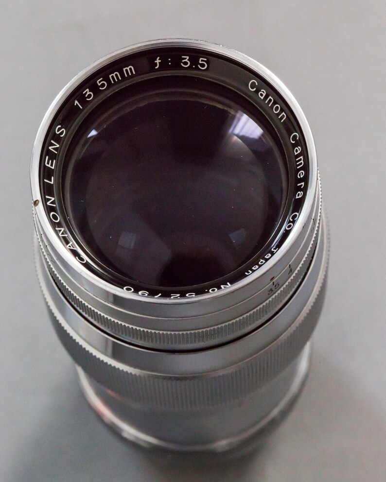 Puede incluir: Un objetivo de c&aacute;mara Canon, 135 mm, f/3.5. El objetivo plateado tiene texto negro, incluyendo "Canon Camera Co." y "Japan". Dise&ntilde;ado para fotograf&iacute;a.