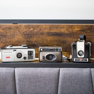 May include: Three vintage cameras arranged in a row. On the left, a white Kodak Brownie Super 27 camera. In the center, a metallic brown Anscomatic 338 camera. On the right, a black and silver Brownie Hawkeye Flash Model box camera with a top handle.