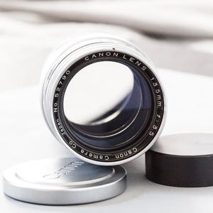 Puede incluir: Un objetivo Canon con cuerpo plateado, texto negro y lente de cristal. El objetivo est&aacute; etiquetado como "Canon Lens 135mm f:3.5" y "Canon Camera Co. Japan No. 52790." Tambi&eacute;n se ven la tapa del objetivo y la tapa trasera.