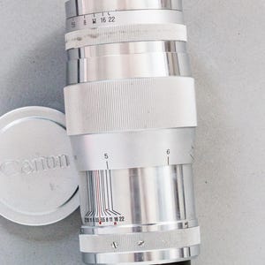 Puede incluir: Un objetivo de c&aacute;mara Canon plateado con base negra y empu&ntilde;adura blanca. El objetivo tiene marcas de medici&oacute;n y una tapa plateada con el logotipo de Canon. Dise&ntilde;ado para fotograf&iacute;a.