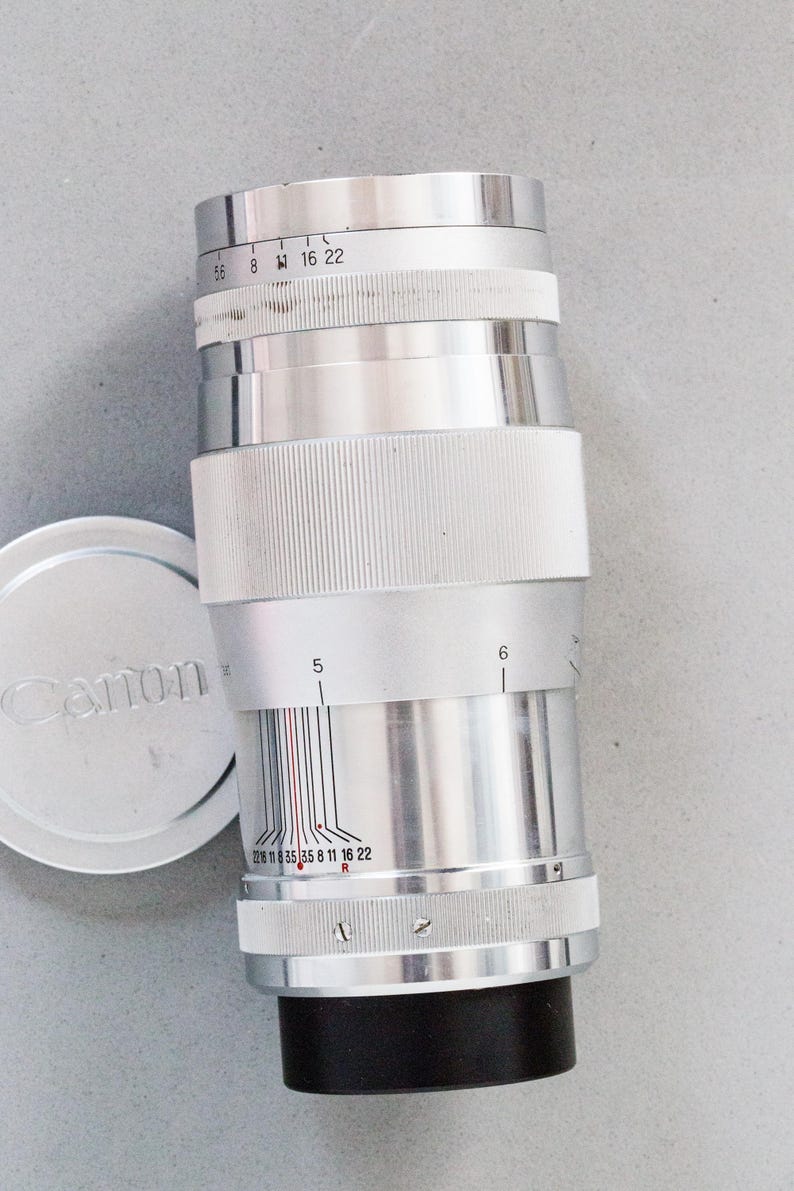 Puede incluir: Un objetivo de c&aacute;mara Canon plateado con base negra y empu&ntilde;adura blanca. El objetivo tiene marcas de medici&oacute;n y una tapa plateada con el logotipo de Canon. Dise&ntilde;ado para fotograf&iacute;a.