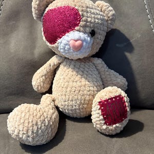 Puede incluir: Un oso de peluche beige hecho a mano con un parche rosa brillante sobre un ojo y una nariz en forma de corazón rosa. El oso tiene un parche rojo en un pie con las letras "AM" cosidas. El oso está sentado sobre una superficie gris.