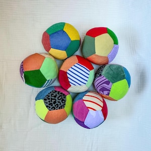 May include: A collection of seven round, multi-colored fabric balls arranged in a cluster. Each ball is constructed from various pentagonal and hexagonal patches featuring diverse patterns, including leopard print, blue stripes, and solid colors.