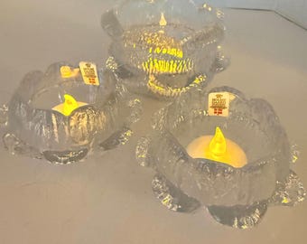 May include: Three clear glass candle holders with a textured, floral design. Each holder contains a yellow LED candle. The candle holders are of varying sizes and are arranged on a white surface.