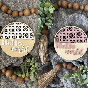May include: Two round wooden signs with a woven pattern on the top half and the words "Hello World" on the bottom half. One sign has a white woven pattern and the other has a pink woven pattern.