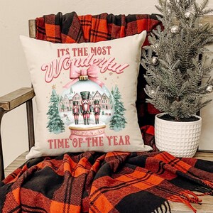 Può includere: Un cuscino decorativo con la scritta "ITS THE MOST Wonderful TIME OF THE YEAR" in corsivo rosa. Il cuscino presenta un design a palla di neve con schiaccianoci e una scena invernale. Il cuscino è su una sedia con una coperta a quadri rossa e nera.