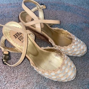 May include: A pair of beige woven wedge sandals with a criss-cross strap and the brand name "ANNA SUI" on the insole.