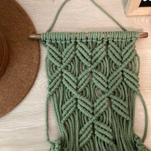 May include: A green macrame wall hanging with a wooden dowel at the top. The hanging has a geometric pattern and is made with thick cotton cord.