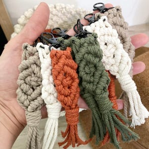 May include: A set of six handmade macrame keychains in beige, white, orange, and green. Each keychain features a unique pattern and a black metal key ring.