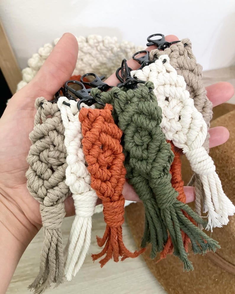 May include: A set of six handmade macrame keychains in beige, white, orange, and green. Each keychain features a unique pattern and a black metal key ring.
