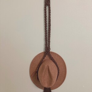 May include: A brown hat hanging on a brown macrame hat holder with a wooden ring at the top.