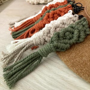 May include: A set of five macrame keychains with tassels in shades of green, brown, and white. The keychains are made with a knotted rope technique and have a metal ring for attaching keys.