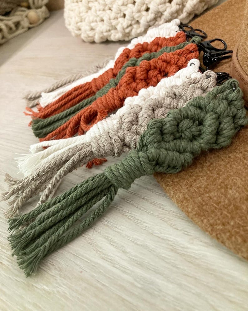 May include: A set of five macrame keychains with tassels in shades of green, brown, and white. The keychains are made with a knotted rope technique and have a metal ring for attaching keys.